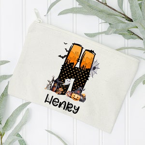 May include: A cream-colored canvas zippered pouch featuring a Halloween-themed design. The design includes a large letter "H" with orange and black accents, a spider web, bats, and the name "Henry" in black script.