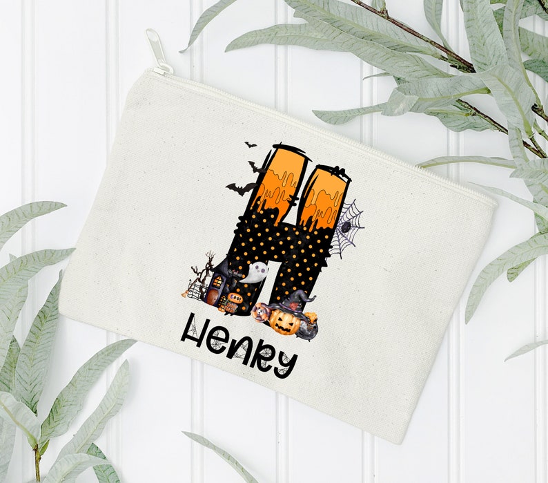 May include: A cream-colored canvas zippered pouch featuring a Halloween-themed design. The design includes a large letter "H" with orange and black accents, a spider web, bats, and the name "Henry" in black script.
