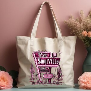 May include: A white canvas tote bag with a pink and white graphic of a retro motel sign that reads "Welcome & Read On Smutville". The sign is surrounded by cacti and stars.