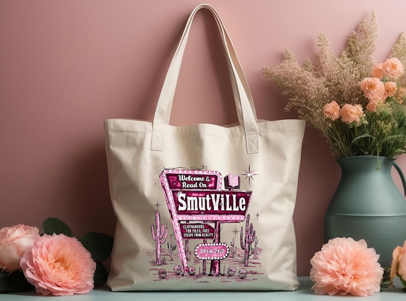 May include: A white canvas tote bag with a pink and white graphic of a retro motel sign that reads "Welcome & Read On Smutville". The sign is surrounded by cacti and stars.
