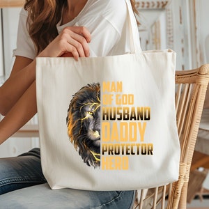 May include: A white canvas tote bag with a black and gold lion graphic and the text "Man of God Husband Daddy Protector Hero".