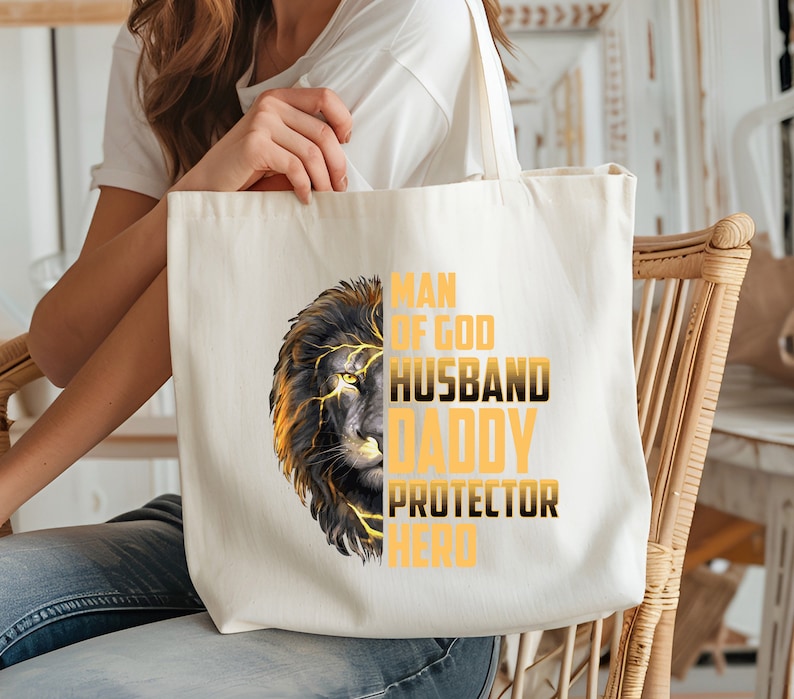 May include: A white canvas tote bag with a black and gold lion graphic and the text "Man of God Husband Daddy Protector Hero".