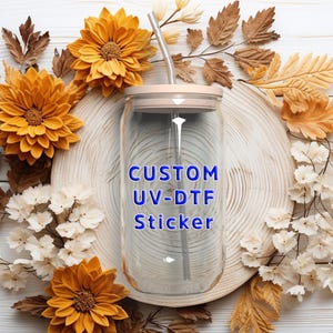 Puede incluir: Un vaso de cristal transparente con tapa de bamb&uacute; y pajita de metal, con el texto azul CUSTOM UV-DTF Sticker impreso en la parte frontal. El vaso tiene una forma cil&iacute;ndrica con la parte superior e inferior ligeramente curvadas.