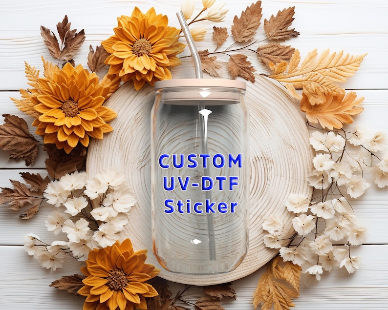 Puede incluir: Un vaso de cristal transparente con tapa de bamb&uacute; y pajita de metal, con el texto azul CUSTOM UV-DTF Sticker impreso en la parte frontal. El vaso tiene una forma cil&iacute;ndrica con la parte superior e inferior ligeramente curvadas.