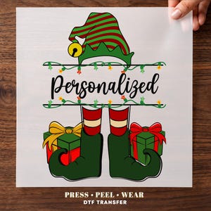 May include: A DTF transfer sheet featuring a graphic of an elf hat with red and green stripes, a yellow bell, and elf legs in green shoes. The design includes a string of Christmas lights, the word Personalized, and two gift boxes with bows.