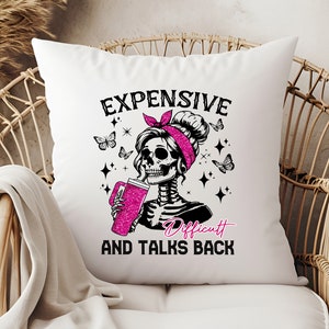 Op de afbeelding: Wit vierkant kussen met een afbeelding van een skeletvrouw met een roze hoofdband, drinkend uit een roze glitterbeker. De tekst luidt "EXPENSIVE AND TALKS BACK" met het woord "Difficult" in roze.