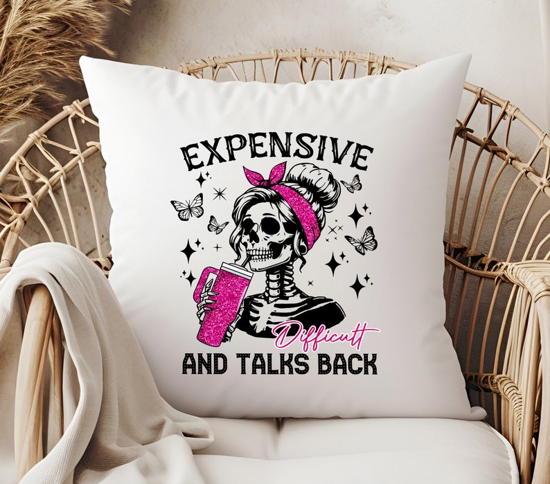 Op de afbeelding: Wit vierkant kussen met een afbeelding van een skeletvrouw met een roze hoofdband, drinkend uit een roze glitterbeker. De tekst luidt "EXPENSIVE AND TALKS BACK" met het woord "Difficult" in roze.