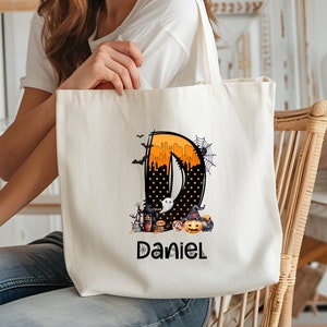 May include: A cream-colored tote bag featuring a Halloween-themed design. The design includes the letter "D" in orange and black, with a spooky illustration of a haunted house, pumpkins, and a ghost. The name "Daniel" is printed below the design.