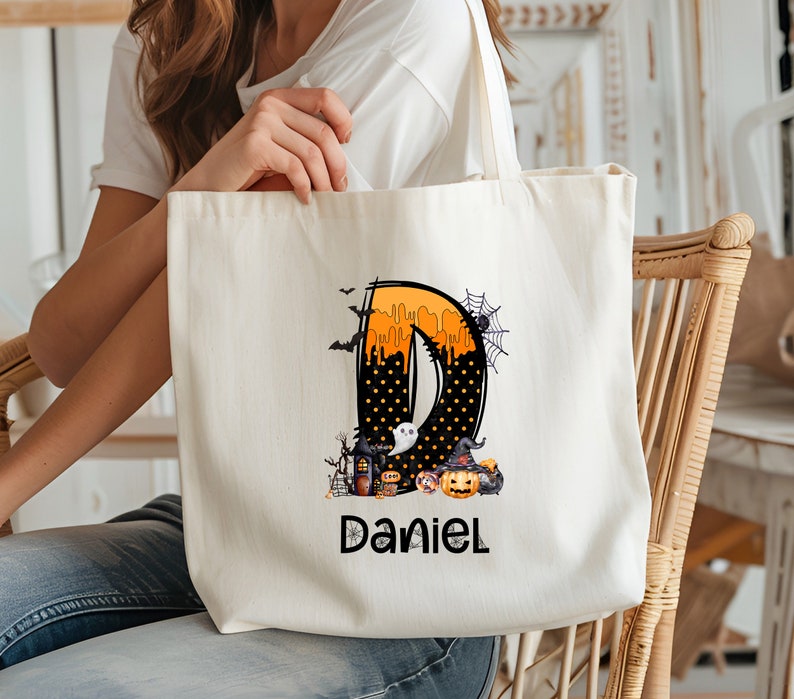 May include: A cream-colored tote bag featuring a Halloween-themed design. The design includes the letter "D" in orange and black, with a spooky illustration of a haunted house, pumpkins, and a ghost. The name "Daniel" is printed below the design.