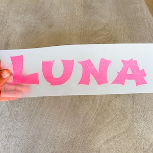 May include: A rectangular sheet of semi-transparent transfer paper featuring the name LUNA printed in large, bold, pink capital letters with a distressed, textured finish. The letters have a stylized, jagged font design.