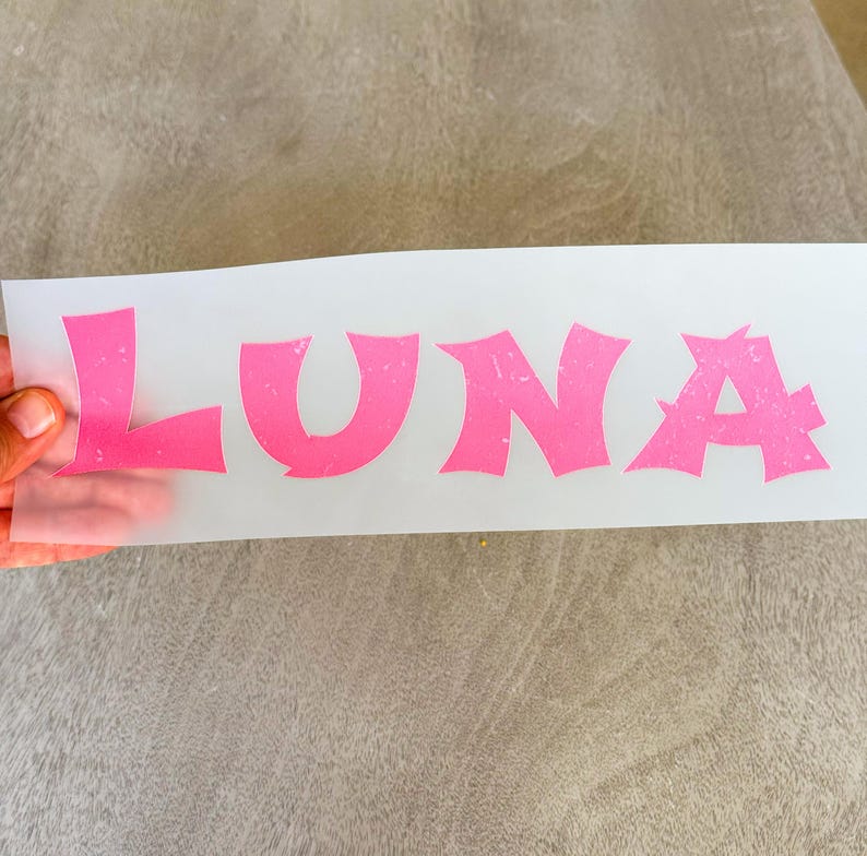 May include: A rectangular sheet of semi-transparent transfer paper featuring the name LUNA printed in large, bold, pink capital letters with a distressed, textured finish. The letters have a stylized, jagged font design.
