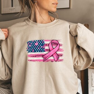 May include: A beige sweatshirt with a pink ribbon and an American flag design. The flag is distressed and has pink hearts and leopard print accents.