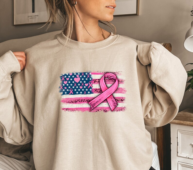 May include: A beige sweatshirt with a pink ribbon and an American flag design. The flag is distressed and has pink hearts and leopard print accents.
