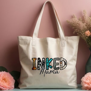以下が含まれることがあります： カラフルな文字で「INKED Mama」と印刷された白いキャンバス地のトートバッグ。文字は、アニマルプリントや花柄など、さまざまなパターンで構成されています。