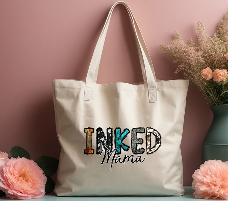 以下が含まれることがあります： カラフルな文字で「INKED Mama」と印刷された白いキャンバス地のトートバッグ。文字は、アニマルプリントや花柄など、さまざまなパターンで構成されています。
