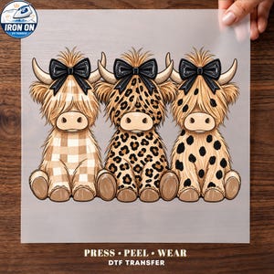May include: A DTF transfer sheet featuring three cartoon Highland cows sitting side-by-side, each wearing a large black bow. The left cow has a beige buffalo plaid pattern, the center has a leopard print, and the right has black spots. Text reads: PRESS PEEL WEAR DTF TRANSFER.