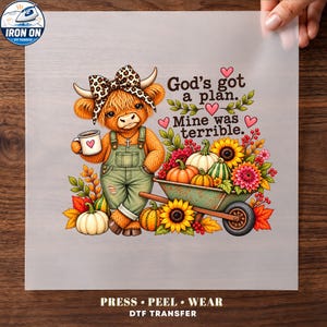 May include: A DTF transfer sheet featuring a cartoon highland cow wearing a leopard print bow and green overalls, holding a coffee mug. The text reads God's got a plan. Mine was terrible. It includes a wheelbarrow filled with pumpkins and sunflowers.