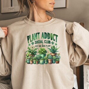 May include: A beige sweatshirt with the text "Plant Addict Social Club" and a graphic of potted plants. The text below the graphic says "Just one more plant."