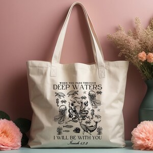 May include: Cream-colored tote bag featuring a black illustration of sea creatures, including an octopus, jellyfish, and whale. The text reads "WHEN YOU PASS THROUGH DEEP WATERS I WILL BE WITH YOU Isaiah 43:2".