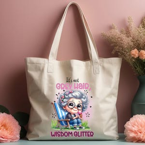 Op de afbeelding: Een witte canvas tote bag met een cartoonillustratie van een vrouw met grijs haar en de tekst "Het is geen grijs haar, het is wijsheid die glinstert".