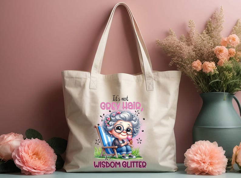 Op de afbeelding: Een witte canvas tote bag met een cartoonillustratie van een vrouw met grijs haar en de tekst "Het is geen grijs haar, het is wijsheid die glinstert".