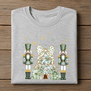 May include: A heather gray t-shirt featuring a graphic print of two green nutcrackers standing on either side of a stylized Christmas tree topped with a large bow. The design includes scattered gold glitter snowflakes and gift boxes at the base.