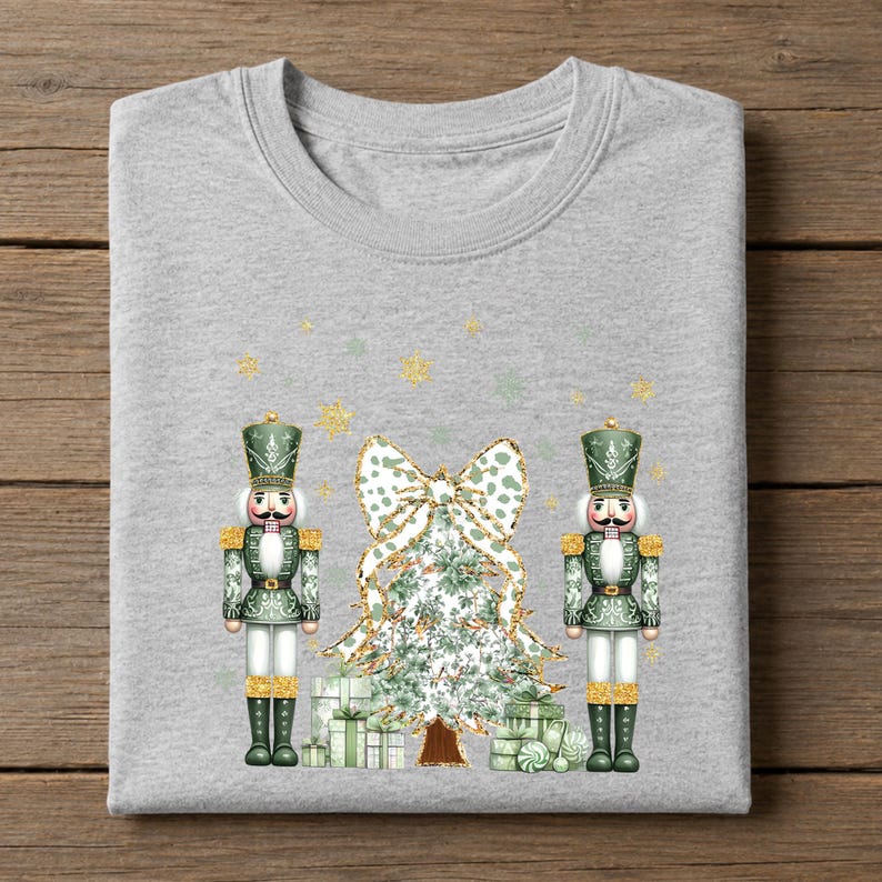 May include: A heather gray t-shirt featuring a graphic print of two green nutcrackers standing on either side of a stylized Christmas tree topped with a large bow. The design includes scattered gold glitter snowflakes and gift boxes at the base.