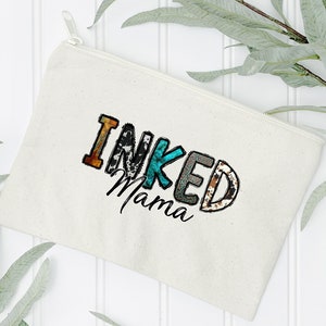 以下が含まれることがあります： パッチワークのデザインでカラフルな文字で「INKED Mama」と書かれた白いキャンバス製のジッパーポーチです。