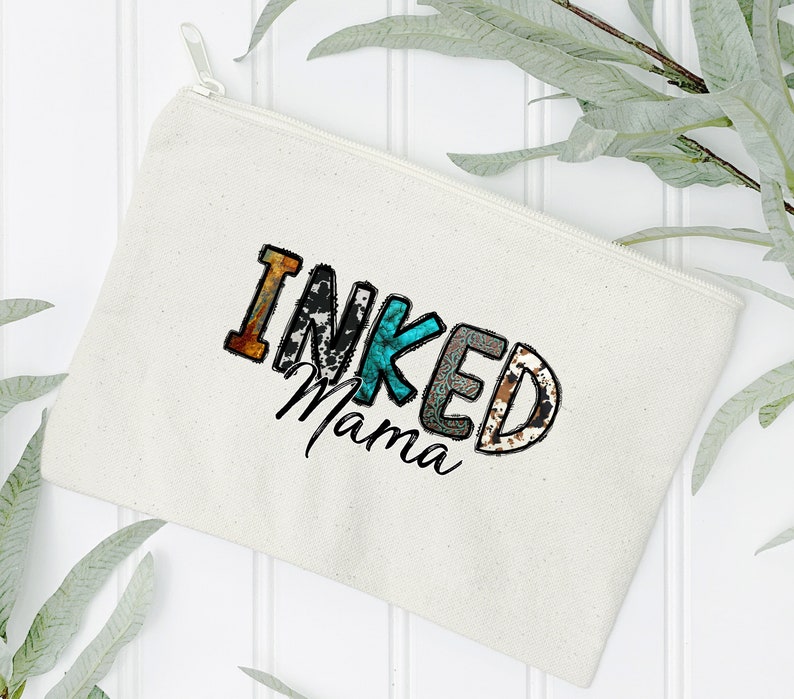 以下が含まれることがあります： パッチワークのデザインでカラフルな文字で「INKED Mama」と書かれた白いキャンバス製のジッパーポーチです。