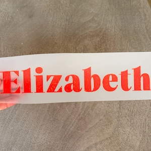 May include: A rectangular, semi-transparent transfer sheet featuring the name Elizabeth printed in a bold, bright orange font with a subtle, textured, speckled appearance throughout the letters, designed for application onto a surface.
