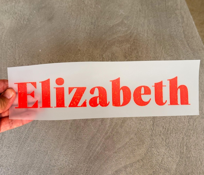 May include: A rectangular, semi-transparent transfer sheet featuring the name Elizabeth printed in a bold, bright orange font with a subtle, textured, speckled appearance throughout the letters, designed for application onto a surface.
