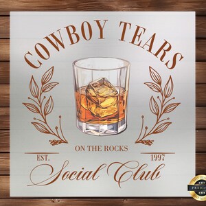 May include: A vintage-style sign with the text "COWBOY TEARS ON THE ROCKS SOCIAL CLUB EST. 1997" in brown lettering on a light gray background. A glass of whiskey with ice is depicted in the center of the sign. The sign has a gold border and a gold seal with the text "BEST QUALITY PREMIUM BEST QUALITY" in the bottom right corner.