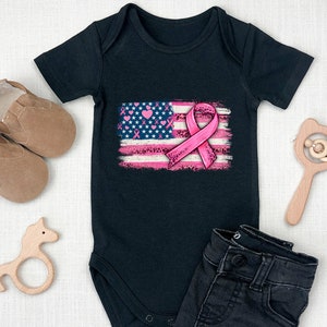 May include: Black baby onesie with a pink ribbon on an American flag design. The flag is distressed and has pink hearts and ribbons on it.