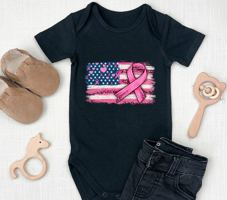 May include: Black baby onesie with a pink ribbon on an American flag design. The flag is distressed and has pink hearts and ribbons on it.