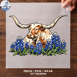 May include: A DTF transfer sheet featuring a graphic illustration of a longhorn cow with brown and white fur, long curved horns, and a tag reading HIGHTFIELD 87. The cow is surrounded by bluebonnet flowers with green leaves and blue petals.