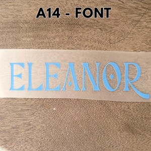 May include: A light blue vinyl decal featuring the name ELEANOR in a stylized serif font with a diamond-shaped accent in the letter O. The decal is presented on a translucent transfer sheet, with the text A14 - FONT displayed above it.