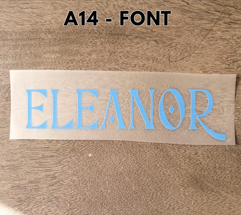 May include: A light blue vinyl decal featuring the name ELEANOR in a stylized serif font with a diamond-shaped accent in the letter O. The decal is presented on a translucent transfer sheet, with the text A14 - FONT displayed above it.