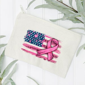 May include: A white canvas zippered pouch with a pink ribbon and an American flag design. The flag is distressed and has pink hearts and ribbons throughout.