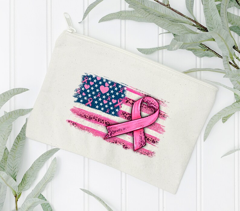 May include: A white canvas zippered pouch with a pink ribbon and an American flag design. The flag is distressed and has pink hearts and ribbons throughout.