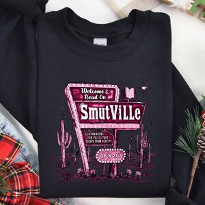 May include: Black sweatshirt with a pink and white graphic of a neon sign that reads "Welcome & Read On Smutville". The sign is surrounded by cacti and has the text "Cliffhangers, Ter Piles, Free Escape From Reality" and "Open 24/7".