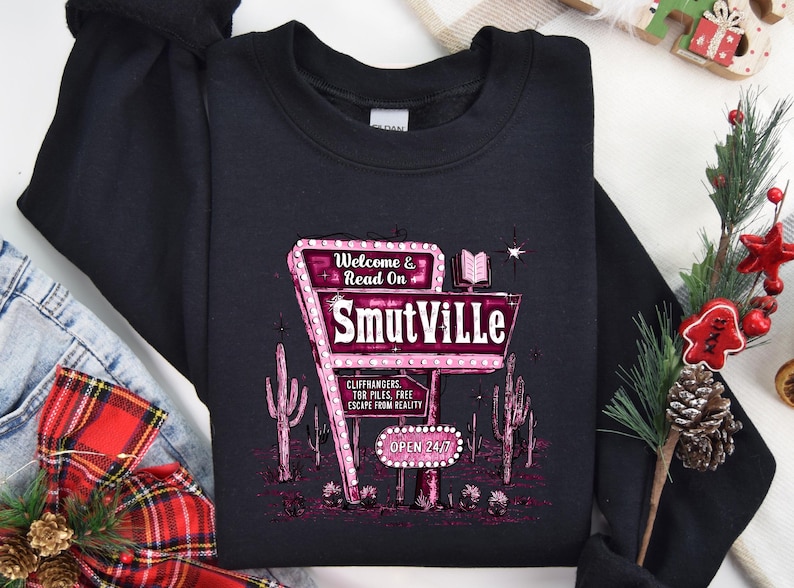 May include: Black sweatshirt with a pink and white graphic of a neon sign that reads "Welcome & Read On Smutville". The sign is surrounded by cacti and has the text "Cliffhangers, Ter Piles, Free Escape From Reality" and "Open 24/7".