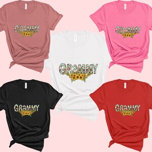 Op de afbeelding: Vijf T-shirts in verschillende kleuren met het woord "Grammy" erop gedrukt in een bloemmotief. De shirts zijn allemaal gevouwen en op een roze achtergrond gelegd.
