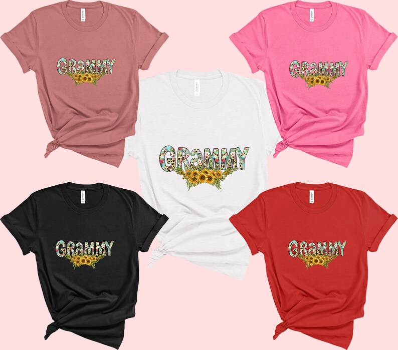 Op de afbeelding: Vijf T-shirts in verschillende kleuren met het woord "Grammy" erop gedrukt in een bloemmotief. De shirts zijn allemaal gevouwen en op een roze achtergrond gelegd.