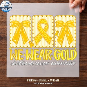May include: A DTF transfer sheet featuring three rectangular panels with yellow bows and a gold awareness ribbon on a striped background. The text reads WE WEAR GOLD childhood cancer awareness in yellow and white lettering with a decorative border.