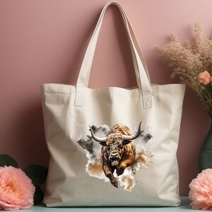 May include: A white canvas tote bag with a brown bull charging through a watercolor-style splash of brown and gray paint.