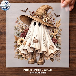 May include: A DTF transfer sheet featuring a ghost wearing a brown witch hat, brown lace-up boots, and patches on its white sheet body. The design is surrounded by autumn-colored flowers, wheat, and small bats. Text at the bottom reads PRESS PEEL WEAR.