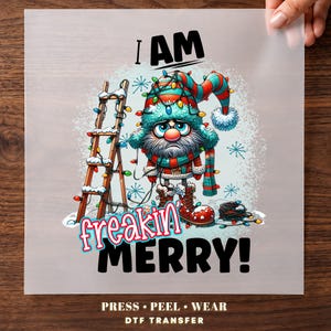 May include: A DTF transfer sheet featuring a cartoon gnome with a long beard, glasses, and a red and teal hat wrapped in colorful Christmas lights. A wooden ladder stands nearby. The text reads I AM freakin' MERRY! and PRESS PEEL WEAR.