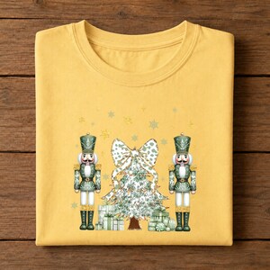 May include: A mustard yellow t-shirt featuring a graphic print of two green and gold nutcrackers standing on either side of a decorated Christmas tree topped with a large bow, surrounded by small gold stars and snowflakes.
