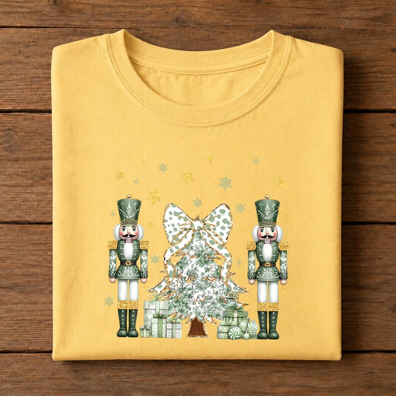 May include: A mustard yellow t-shirt featuring a graphic print of two green and gold nutcrackers standing on either side of a decorated Christmas tree topped with a large bow, surrounded by small gold stars and snowflakes.