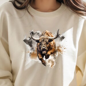 May include: A white sweatshirt with a brown and black bull illustration. The bull is charging forward with its mouth open and horns pointed forward. The bull is surrounded by a watercolor-style background of brown and gray splatters.